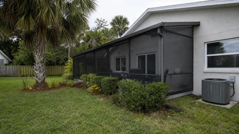 Gray stucco home with screened patio enclosure, lawn, shrubs, and palm trees